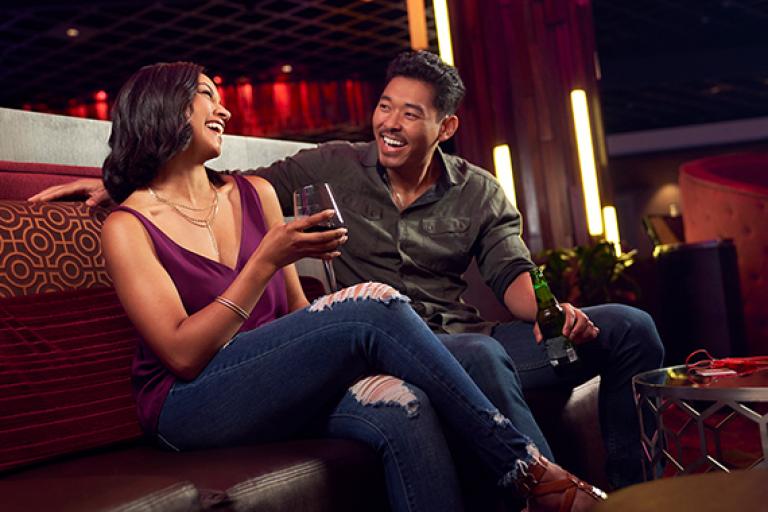 Two young adults laugh while sipping on beer and wine on a large upholstered bench 