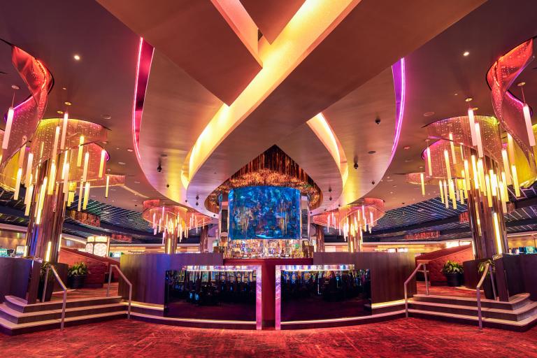 Luxurious casino interior with vibrant pink and purple lighting, featuring a bar at the center under a large x-shaped ceiling design, surrounded by decorative vertical light fixtures.
