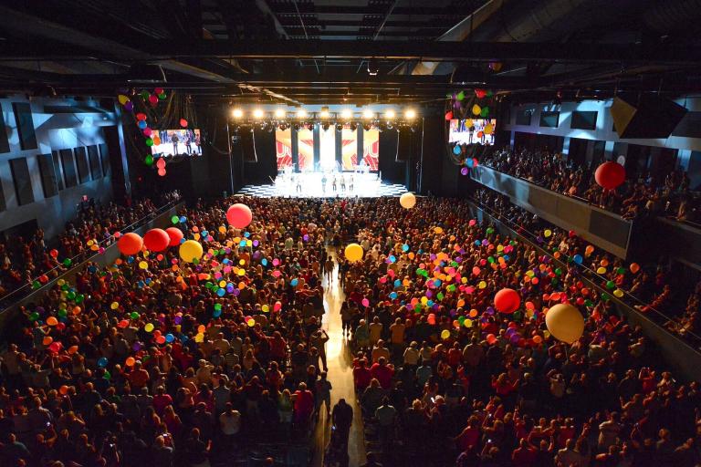 An indoor concert venue filled with people holding colorful balloons, focusing on a brightly lit stage with performers.