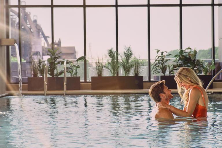 A couple embraces in an indoor swimming pool, bathed in sunlight streaming through large windows. the serene setting includes decorative plants and a spa-like ambiance.