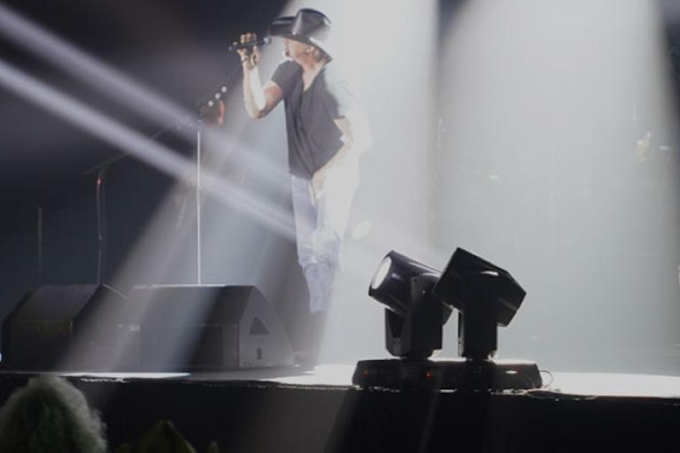 A singer in a hat performing onstage under bright spotlight beams, with stage equipment visible in the foreground and the audience blurred in the front.