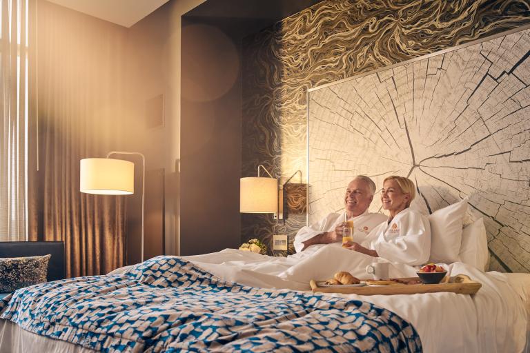 A mature couple enjoys breakfast in bed in a luxurious hotel room, bathed in warm sunlight, smiling and toasting with orange juice.