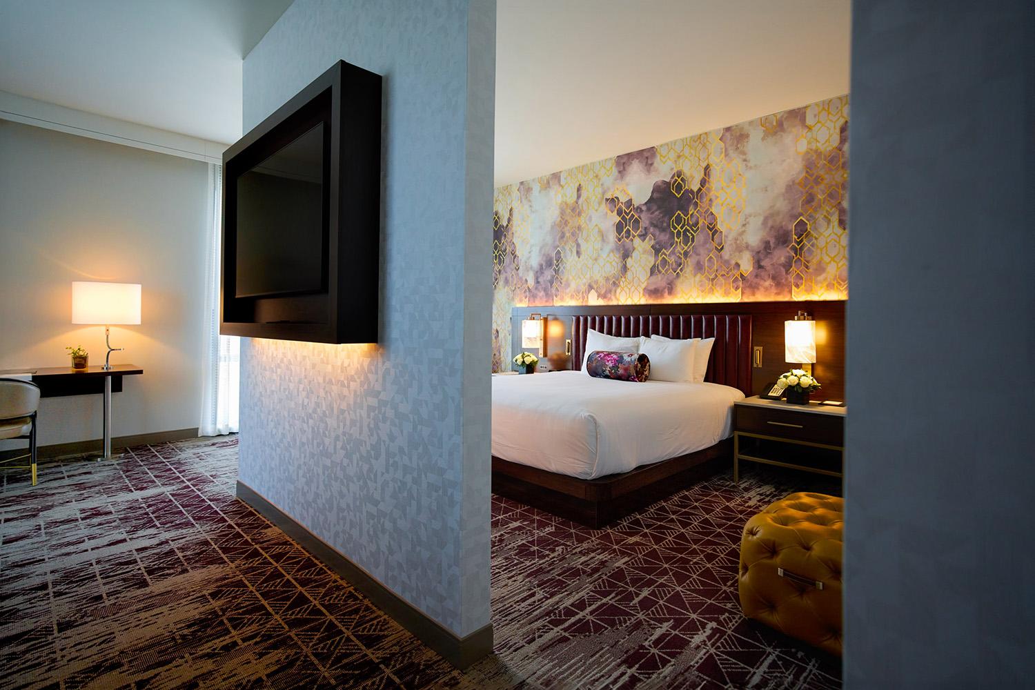 A modern hotel room with a king bed, flat screen TV, geometric carpeting and a yellow ottoman