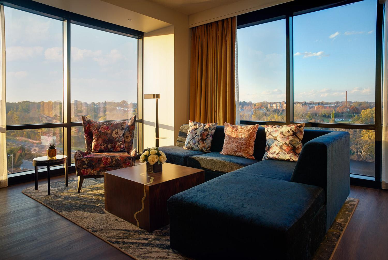 A modern hotel room with a large sectional, a wing-backed chair and reclaimed wood coffee table