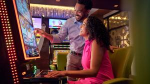 A man stands next to a seated woman at a slot machine, both are smiling