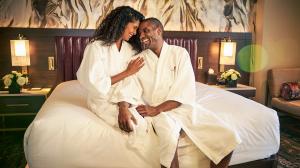 A joyful couple in white robes sits closely on a bed in a luxurious hotel room, laughing together with an abstract art piece above the bed and subtle lighting.