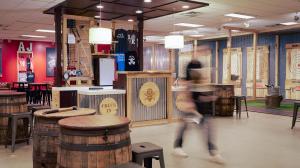 Indoor axe throwing venue with a rustic design featuring barrel tables, hanging lamps, and a check-in counter. a blurred figure walks by, adding motion to the scene.