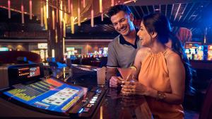 A joyful couple enjoys drinks while playing at a video poker machine in a vibrant casino setting with colorful lights and gaming machines in the background.