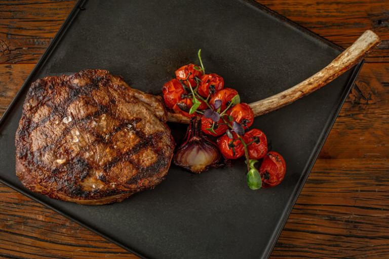 A grilled tomahawk steak with charred exterior and roasted cherry tomatoes on a black plate.