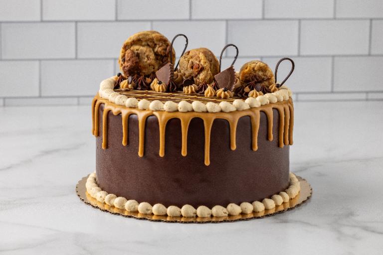 A chocolate cake with caramel drizzle and chocolate chip cookies on top, presented on a white marble surface.