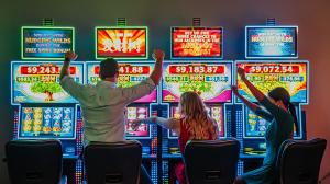 Three people sitting at bright slot machines in a casino, excitedly interacting with the games which display colorful lights and jackpot amounts.