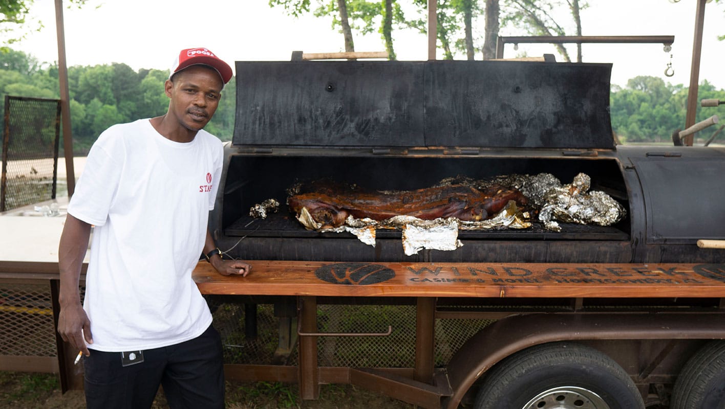 Wind Creek employee standing in front of a large smoker cooking pork. The meat is covered in sauce and had been wrapped in foil.