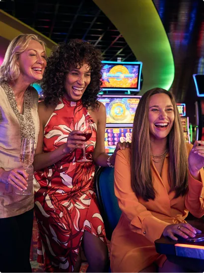 A woman plays a slot machine while 2 others cheer her on.