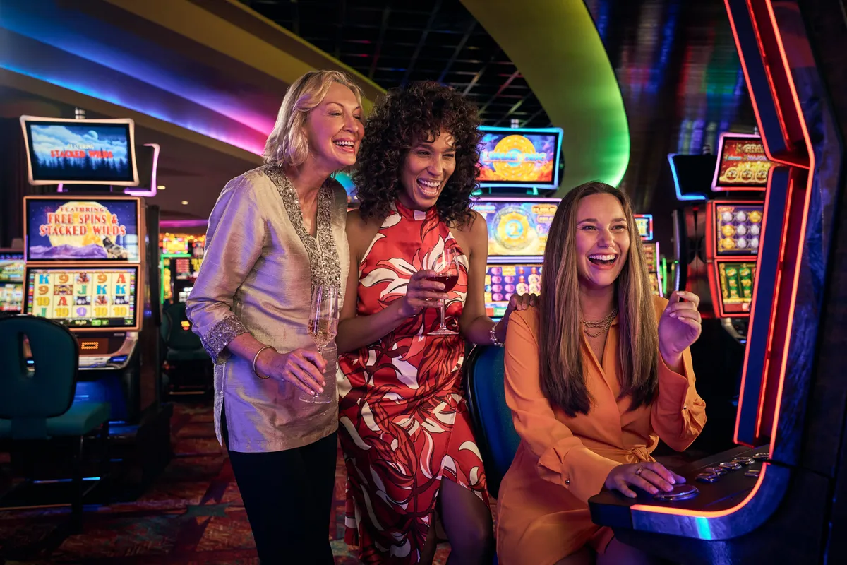 A woman plays a slot machine while 2 others cheer her on.