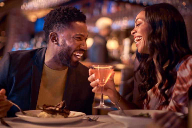 A man and a woman laughing while enjoying cocktails and a meal