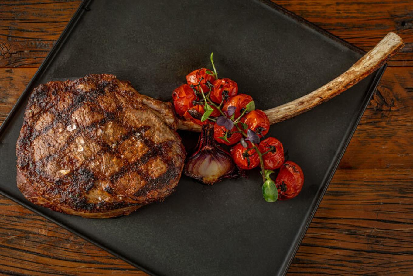 A grilled tomahawk steak with charred exterior and roasted cherry tomatoes on a black plate.