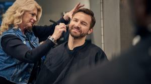 A stylist performs a beard trim on a man wearing a salon cape